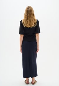 Črn top s kratkimi rokavi in dolga mornarsko modra krila, gladka tekstura, do kolen z razporkom na hrbtni strani. Svetlo rjava obutev za na svinčnik, model obrnjena proti hrbtu.