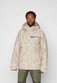 Veste à capuche en camouflage beige avec poche zippée, tissu texturé et cordons de serrage. Assortie à un pantalon vert.