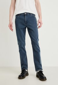 Calças de ganga azul para homem com um design de perna reta, estilo de cinco bolsos, desbotamento subtil e fecho de botão. Combinadas com sapatos pretos.