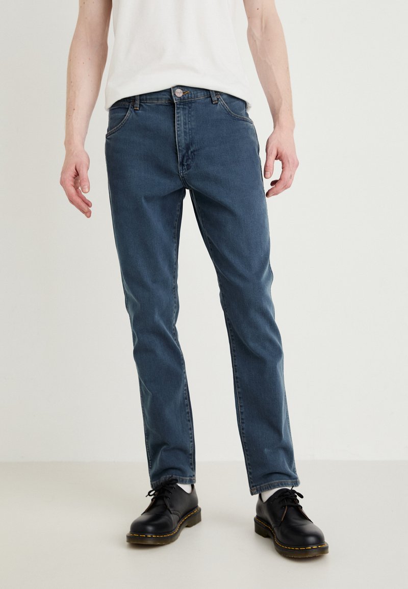 Calças de ganga azul para homem com um design de perna reta, estilo de cinco bolsos, desbotamento subtil e fecho de botão. Combinadas com sapatos pretos.