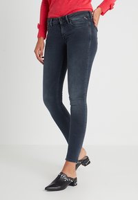 Jeans ajustados de mezclilla en un lavado oscuro con una textura suave, que presentan una cintura de altura media, un ajuste delgado y detalles de costura sutiles. Combinados con zuecos negros.