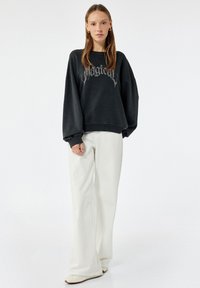 Czarny oversize sweter z napisem "Magical", z prążkowanymi mankietami, w połączeniu z luźnymi białymi spodniami i spiczastymi beżowymi butami, na jasnym tle.