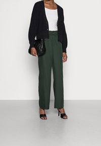 Cardigan en tricot noir sur un débardeur blanc, associé à un pantalon vert taille haute et des sandales à talons noires. Sac en cuir matelassé noir tenu à la main.