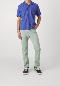 Kortärmad blå skjorta med två bröstfickor, parat med ljusgröna byxor och svarta sneakers med vit sula och gröna detaljer.