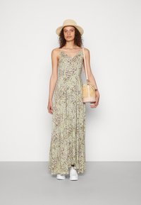 Florales Maxikleid in Hellgrün mit Spaghettiträgern, ausgestattet mit einem V-Ausschnitt und weißen Akzent-Schuhen; trägt eine runde, geflochtene Handtasche.