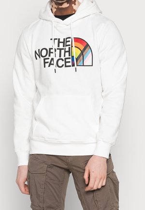 Henkilö pukeutuneena valkoiseen huppariin, jossa on monivärinen sateenkaarilogolla varustettu The North Face -merkki, yhdistettynä ruskeisiin cargo-housuihin, näkyvissä niskasta reisien keskivaiheille saakka.