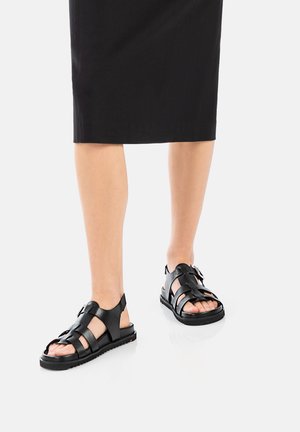 Black leather sandals featuring a strappy design, open toe, adjustable buckle, and textured sole for grip, worn with a black skirt.