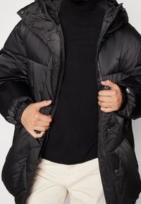 Svart puffjacka med huva, med dragkedja och tryckknappar, elastiska ärmslut och en slät, glänsande yta. Buren  över en svart tröja.