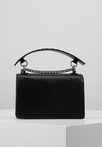 Borsa di pelle nera con una catena spessa e un manico largo in pelle, posizionata su un piedistallo bianco contro uno sfondo grigio uniforme.