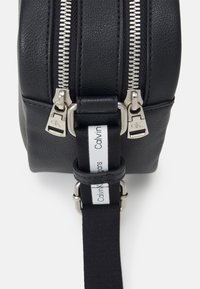 Černá kožená taška s dvojitými stříbrnými zipy, texturovaným povrchem a černým popruhem s bílým logem Calvin Klein.