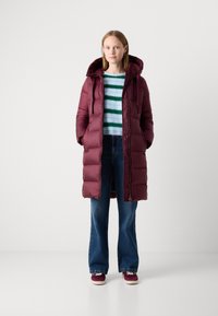 Bordeaux pufferjack met een capuchon, met een gewatteerd ontwerp. Gecombineerd met een gestreepte trui, uitlopende jeans en bordeauxkleurige schoenen.