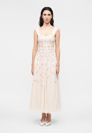 Kvinde står med fronten mod kameraet iført en ærmeløs, flared cremegul kjole med pink blomsterbroderi og hvide spidse sko.