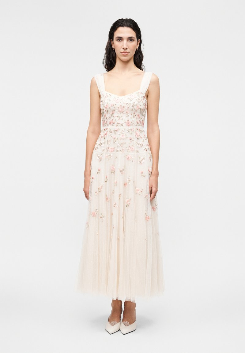 Γυναίκα στέκεται προς τα εμπρός φορώντας αμάνικο, εκρού φόρεμα μέχρι τον αστράγαλο με ροζ κεντήματα λουλουδιών και λευκά μυτερά παπούτσια.