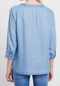 Ljust blå denimskjorta med trekvartslånga ärmar, draperad halsringning och knäppta manschetter. Slät textur och avslappnad passform.