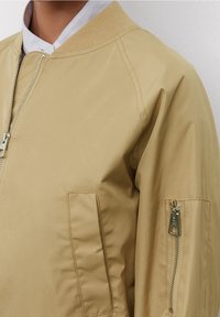 Giubbotto bomber khaki con zip frontale, colletto e polsini a costine, tasca sul braccio sinistro con zip e una texture liscia e leggermente lucida.