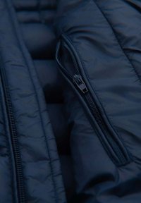 Schwarze gepolsterte Jacke mit glatter Textur. Verfügt über einen sichtbaren Reißverschluss an der Seite und eine gesteppten Detail entlang der Stoffoberfläche.