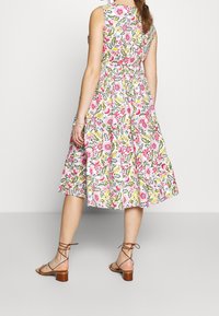 Vestido midi floral con un fondo blanco y patrones multicolores, tiene un corsé ajustado, cinturilla con lazo y una falda fluida. Sandalias marrones con tiras.