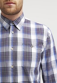 Napapaidan kauluspaidassa on ruudullinen kuvio sinisen, harmaan ja valkoisen sävyissä. Se on valmistettu kudotusta kankaasta ja siinä on rintatasku sekä kaulussuunnittelu.