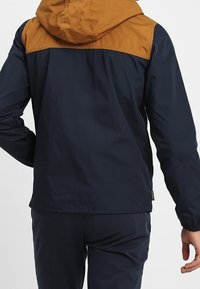 Giacca blu navy con cappuccio e yoke marrone. Presenta un design semplice, polsini elasticizzati e una linea dritta. Tessuto strutturato visibile.