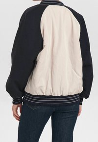 Veste bomber bleu marine et crème, avec un design matelassé, des poignets côtelés et un ourlet élastiqué avec des rayures blanches. Tissu lisse et texturé.