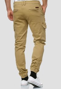 Beige cargo pants with elastic cuffs, side pockets, and patch details. Constructed from durable fabric for a relaxed fit.