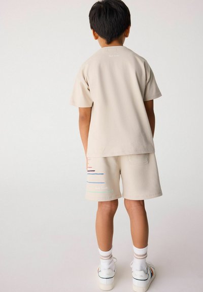 Beige bomull t-shirt och shorts-set med flerfärgade horisontella ränder. Enfärgad baksida, med varumärkeslogga på t-shirten. Vita strumpor och skor.