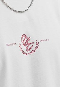 Λευκό μπλουζάκι με κόκκινο κείμενο "Creative Dept. Copenhagen", περίτεχνα αρχικά "CRG" και σχέδιο δάφνινου στεφανιού κάτω από ασημένιο κολιέ με αλυσίδα.