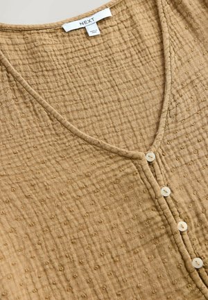 Beige, strukturierte Bluse mit V-Ausschnitt und kleinen Knöpfen auf der Vorderseite. Sie besteht aus einem weichen, zerknitterten Stoff mit punktuellen Mustern.