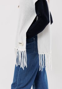 Poncho bianco con frange realizzato in materiale morbido; presenta un design aperto e un contrasto di colore con maniche blu scuro. Etichetta sul lato.
