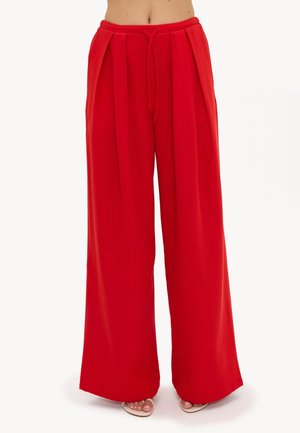 Pantalon rouge ample taille haute avec plis sur le devant et taille à cordon, porté avec des sandales ouvertes beiges.