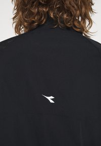 Svart jacka med en mjuk textur, med en vit logotypaccent på baksidan och en modern, minimalistisk design. Ingen synlig hårdvara.