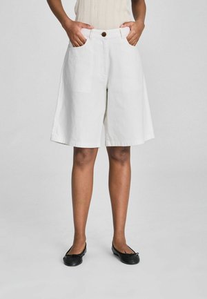 Osoba nosząca białe szerokie szorty do kolan z przednimi kieszeniami, beżową bezrękawnik i czarne płaskie buty, stojąca z rękami w kieszeniach.