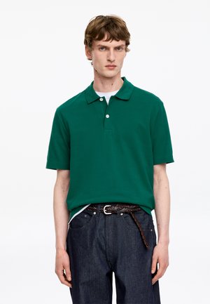 Jeune homme portant un polo vert à manches courtes, un jean foncé et une ceinture tressée marron, debout devant un fond blanc.