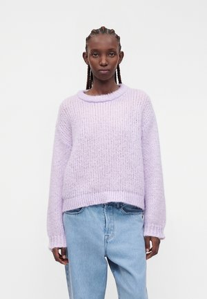 Adulte portant un pull en maille violet clair et un jean bleu clair, debout devant un fond uni gris clair, face à l’avant.