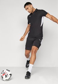 Svart polo-skjorte med korte ermer og hvite sidedele, kombinert med svarte shorts. Svarte fotballsko og hvite sokker. Fotball på gulvet.
