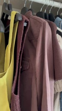 Top giallo senza maniche, gilet di maglia marrone, giacca malva e altri vestiti assortiti appesi a grucce nere in un armadio.