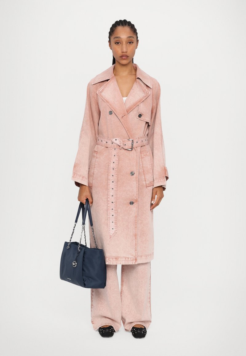Trench coat rosa chiaro in stile denim con bottoni doppi, cintura e applicazioni laterali. Abbinato a pantaloni a gamba larga e una borsa blu navy.