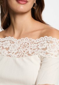 Cremefarbenes Off-Shoulder-Top aus strukturiertem Rippstoff mit weißen Spitzenbesätzen, die florale Muster entlang des Ausschnitts aufweisen.