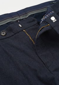 Donkerblauwe broek met een gestructureerde stof, gouden rits, zwarte knoop en een patronen binnenband. Gedetailleerde stiknaden zichtbaar.