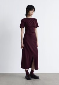 Burgunderfarbenes Midi-Kleid mit kurzen Ärmeln, drapiertem Design und seitlichem Schlitz, aus glattem Stoff; kombiniert mit dunklen Loafers und weinroten Socken.