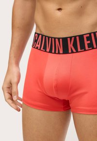 Calvin Klein muške gaće u svijetlo koraljnoj boji, izrađene od rastezljivog materijala. Imaju crni pojas s istaknutim logotipom branda u crvenoj boji.