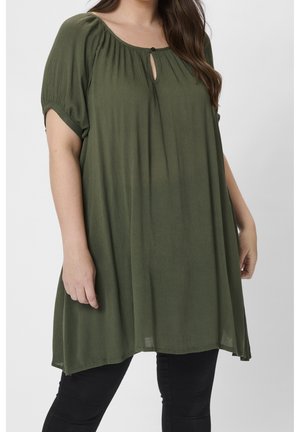 Femme portant une tunique ample vert olive à manches courtes et encolure en trou de serrure, associée à un pantalon noir, debout devant un fond uni.