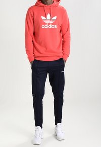 Felpa rossa in cotone con logo bianco Adidas, abbinata a pantaloni da ginnastica blu con strisce bianche e scarpe sportive bianche.