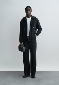 Mladý muž stojaci, oblečený v čiernej mikine na zips, bielom tričku, čiernych nohaviciach a čiernych topánkach, držiaci v pravej ruke malú tmavosivú tašku.