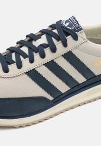 Beige Leder-Sneaker mit marineblauen Lederakzenten, drei marineblauen Streifen und einer strukturierten Zehenkappe. Verfügt über flache Schnürsenkel und eine gepolsterte Sohle.