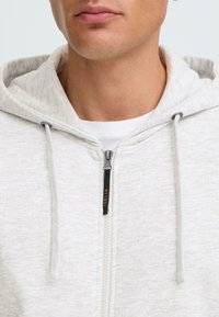 Svijetlosiva zip-up majica s mekanom teksturom, podesivim kapuljačom i crnim patentnim zatvaračem s narančastim detaljem brendiranja.