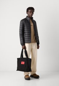 Zwarte pufferjack met horizontale stiksels, beige sweatshirt, crèmekleurige broek en zwarte totebag met rode label; bruine instappers.