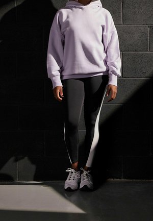 Světle fialový oversized mikina, černé legíny s bílým postranním panelem a bílé sportovní boty. Měkká látka, uvolněný střih a moderní design.