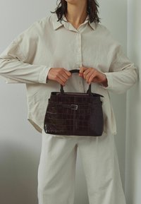 Geantă de mână de culoare brown închis, cu model de crocodil, cu o formă structurată, mânere duble și închidere cu fermoar. Modelul poartă o cămașă supradimensionată de culoare bej deschis.