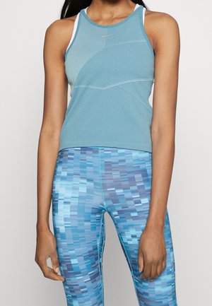 Kobieta w niebieskim sportowym topie bez rękawów i niebieskich legginsach w pikselowy wzór, stojąca na białym tle.